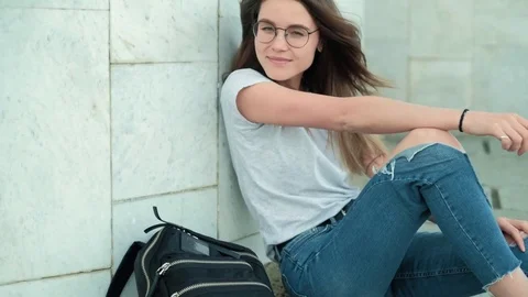 Girl brunette in glasses with a black backpack sits on a gray marble wall Stock Footage