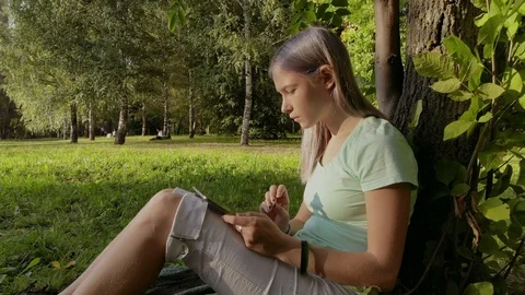 Girl checking email with ipad sitting under a tree on the grass in the park Stock Footage 100613397