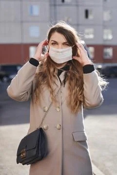 Girl in a coat and with a bag on a sunny street puts on her medical mask Stock Photos