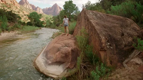 Girl crosses large boulder while river flows towards distant mountains Stock Footage 321428550