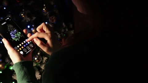 Girl decorating Christmas tree using her phone. Uses smart home app 스톡 동영상 287858414