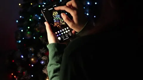 Girl decorating Christmas tree using her phone. Uses smart home app 스톡 동영상 287858419