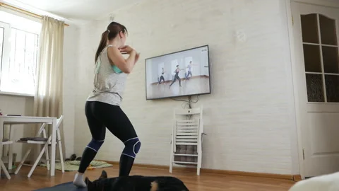 The girl does exercises while watching a program on TV at home. A young woman in Stock-Footage 129705593