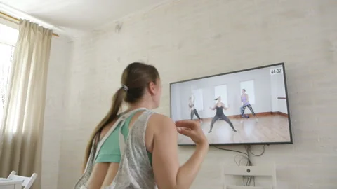 The girl does exercises while watching a program on TV at home. A young woman in Stock Footage 129908859