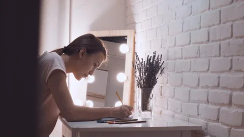Girl draws in a notebook. In the background lights, make-up mirror. Brick wall Stock Footage 101290345