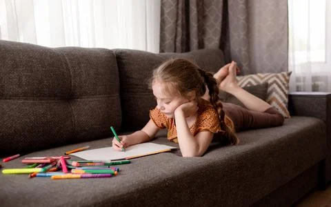 The girl draws while lying on the couch in the living room. The child spends her Stock Photos