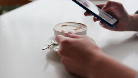 A girl drinks coffee sits in a cafe looks at the smartphone screen choosing Stock Footage 224716592