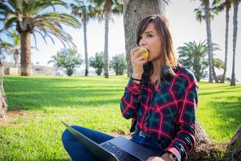 Girl eats an apple while using the laptop in a park in the open air. Stock Photos