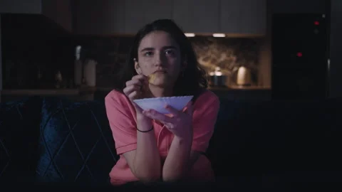 Girl eats crisps while sitting on the sofa in front of the TV at night Stock Footage 324089668