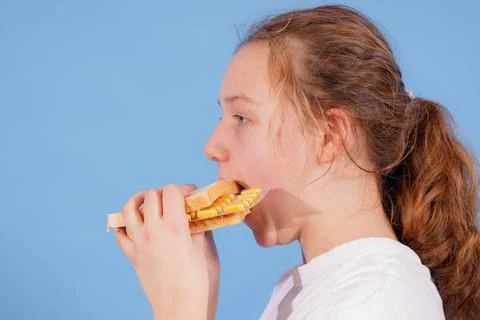Girl eats sandwich while sitting against a blue background during daytime 库存照片