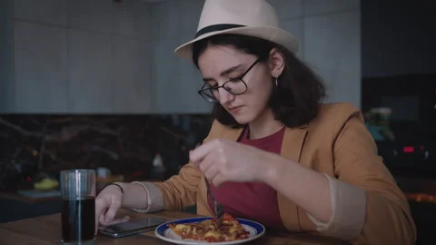 Girl Eats a Slice of Pizza While Sitting at a Table Wearing a Hat and Browsing Stock-Footage 315999942