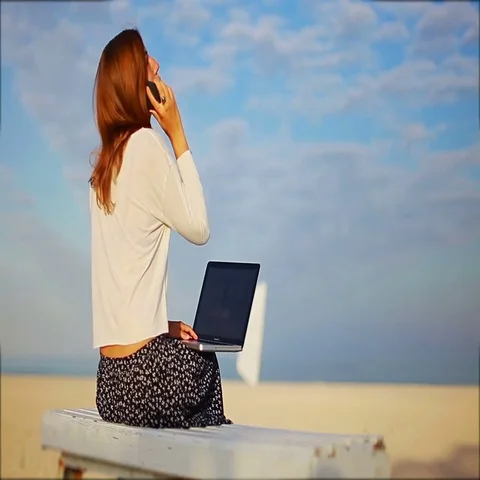 Girl ends a phone conversation while working with a laptop on the beach Stock Footage 69465207