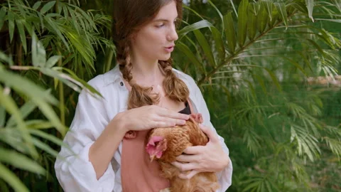 Girl gently strokes a rooster while surrounded by lush greenery on an eco farm Stock Footage 301517909