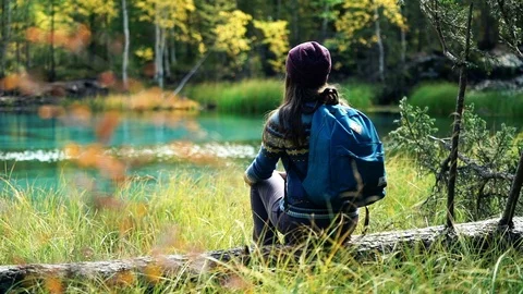 Girl in hat with backpack sits on a fallen tree by blue mountain lake. Back view 库存影片 97408165