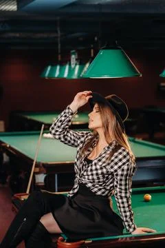 A girl in a hat in a billiard club sits on a billiard table.Playing pool Stock Photos