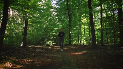 Girl jogging through a sunlit forest trail, surrounded by lush green trees .. Stock Footage 307828126
