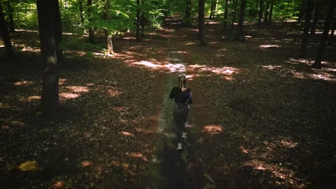 Girl jogging through a sunlit forest trail, surrounded by lush green trees .. Stock Footage 307828162
