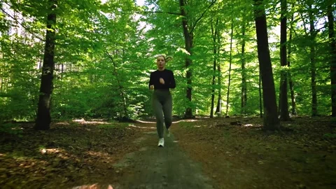 Girl jogging through a sunlit forest trail, surrounded by lush green trees .. Stock Footage 307828173