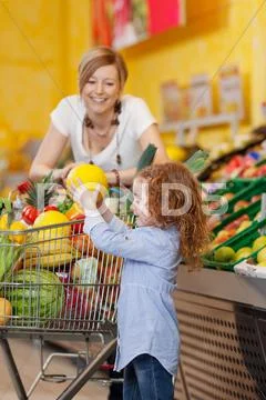Girl keeping muskmelon in shopping cart while mother looking at Stock Photos