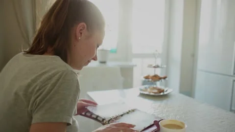 Girl kitchen opposite the window, at the table, leafs through a notebook Stock Footage 150437290
