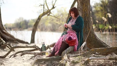 A girl knits clothes from wool while sitting on the roots of a tree Stock Footage 309233332