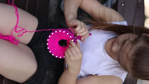 A girl knits on a loom with pink threads while sitting on a bench in a summer Stock Footage 280306352