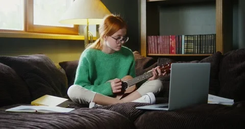 A girl learning to play ukulele while watching video tutorial on laptop Stock Footage 164054773