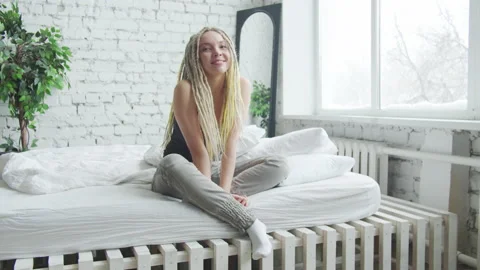 A girl with light dreadlocks sits on a bed with white sheets. She smiles and Stock Footage 156209982
