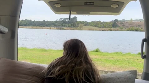 A girl looking out from the back of a camper van towards a lake at Horahora Stock Footage 320873107
