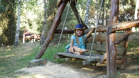 Girl looks into the camera while sitting on a wide wooden swing. Video stock 93669011