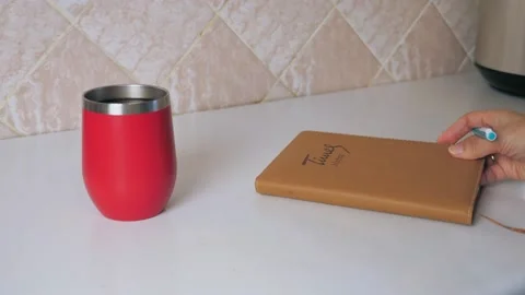 The girl makes a note in a notebook at the kitchen table and drinking hot coffee Stock Footage 214355433