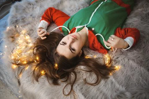 A girl in a New Year's overalls with a festive mood lies on a bed around lumi Stock Photos
