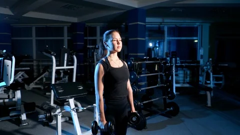 The girl performs an exercise lifting hands with dumbbells in front of you. 4k Stock Footage 99727098