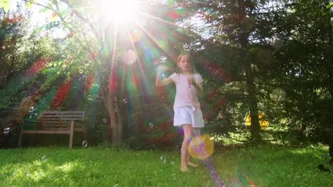 Girl playing with soap bubbles while her dog runs in a sunlit garden Stock Footage 299710498
