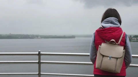Girl pulls hood of her jacket over her head in drizzling rain next to river. Video stock 134697790