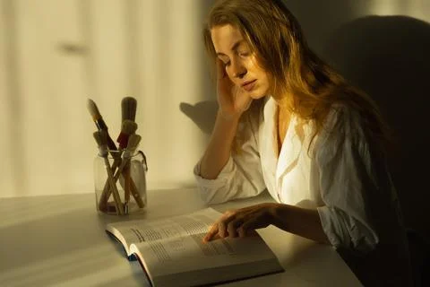 A girl reading a book in the room at the table, in the sunlight. Spare time Stock Photos