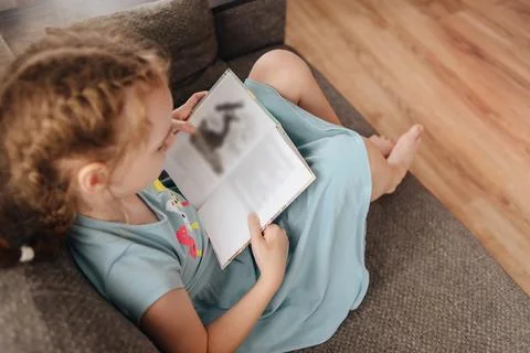 Girl reading a book for school program sitting on the couch. The theme of books Stock Photos