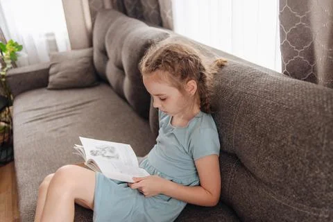 Girl reading a book for school program sitting on the couch. The theme of books Stock Photos