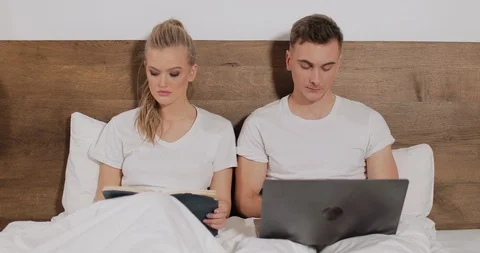 Girl Reading Book while Man using Laptop in Bed Видео 103827751