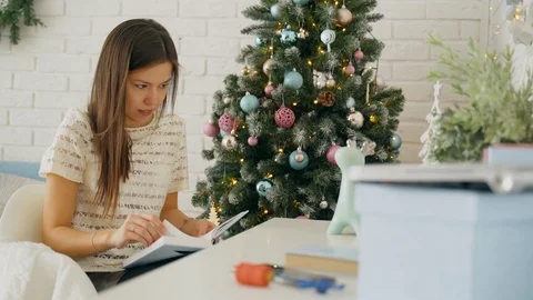 A girl is reading a book while sitting at a table near the Christmas tree. Stock Footage 103532995