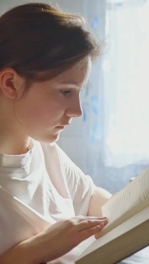 Girl reading a book while sitting at a table near a window. Vertical view Stock Footage 276483046