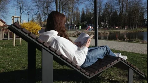 A girl reading a novel while sitting on a deck chair by a pond. Stock Footage 305761666
