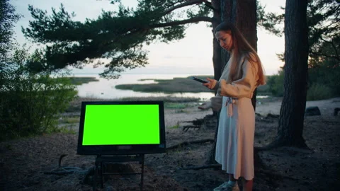 Girl with a remote control in her hand hits a TV with a green screen in forest 库存影片 253178257