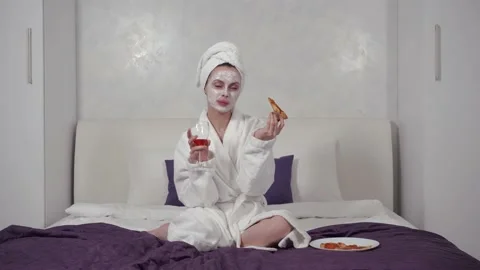 A girl in a robe appetizingly eats pizza and drinks wine sitting on the bed Stock Footage 153543338