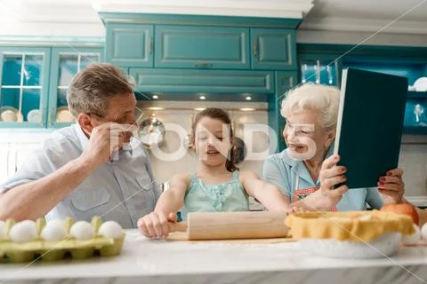 Girl rolling dought out, sitting at the kitchen table with joyful grandparents Stock Photos