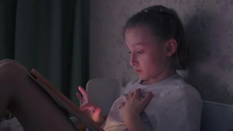 Girl scrolls tablet in dim bedroom at bedtime, blue screen glow on face, quiet Stock Footage 331987452