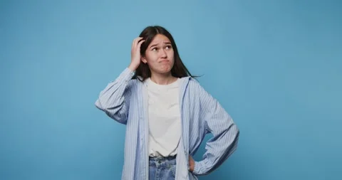 Girl showing confusion while scratching her head against a blue background .. Stock Footage 313273671