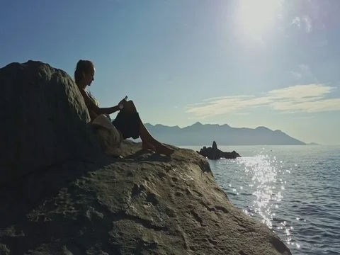 Girl Sits by Bag on Stone Listens to Music against Ocean Stock Footage