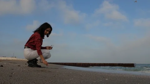 Girl sits on the beach and throws pebbles at sea against the sky Stock Footage 102016283