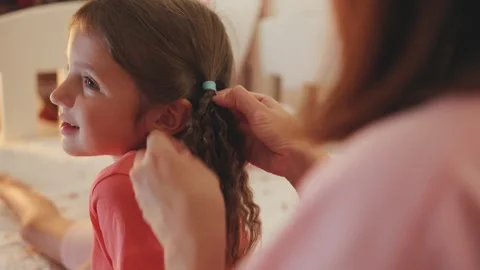 Girl sits on bed while her mother styles her hair in a warm and cozy room during Stock Footage 330749195